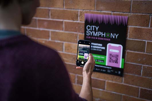 A person scanning a City Symphony poster QR code with their phone