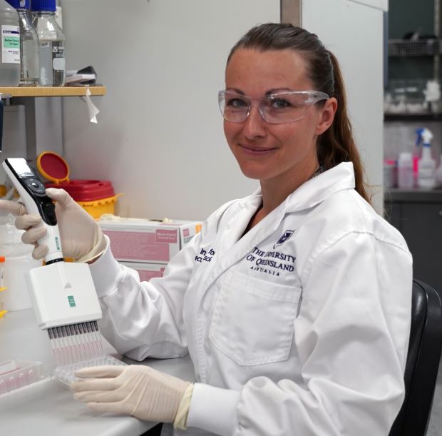 A woman in a white lab coat, goggles and gloves holds a piece of equipment in a laboratory, she is smiling at the camera