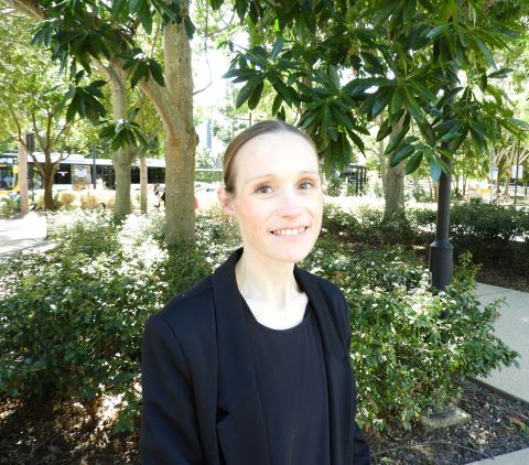 A white woman stands outdoors with trees and a bus behind her. She has her hair pulled back and is smiling.