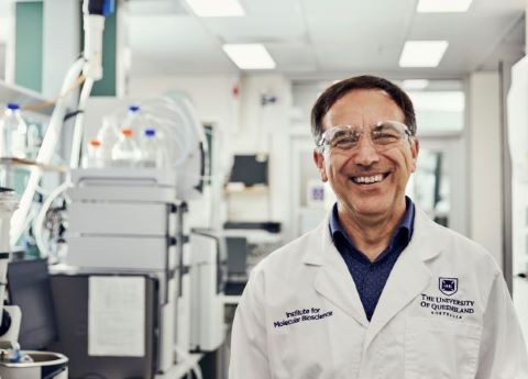 A scientist in white coat and protective goggles stands in a laboratory, smiling broadly.