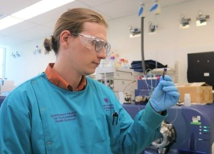 A male scientist in goggles and lab coat stands in a laboratory looking at a test tube