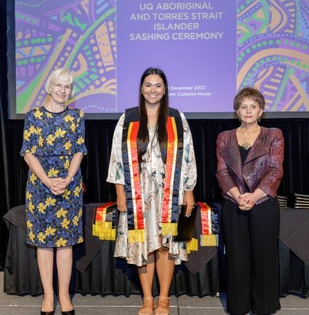 Three women stand on a stage in front of a sign reading UQ Aboriginal and Torres Strait Islander sashing ceremony. The young woman in the centre is wearing a black, red and yellow sash and is smiling broadly.