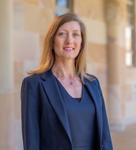 A woman with shoulder length brown blonde hair smiles at the camera. She is wearing a navy top and jacket and a pendant necklace, and is standing with the UQ sandstone cloisters in the background.