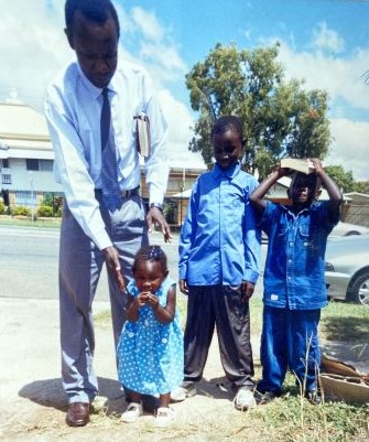 An African family stands outside dressed up - a father is wrangling a young girl while her brothers watch on.