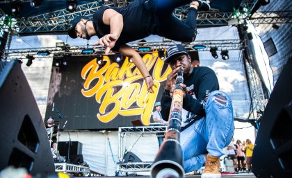 Baker Boy on stage. One performer is captured mid-flip, while another plays the didgeridoo, with the end of it pointing towards the camera.