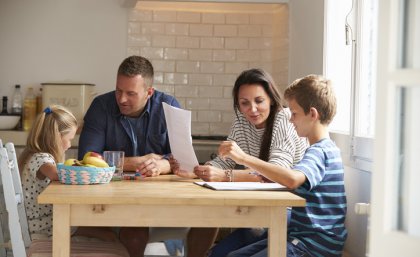 A mother and father sit at the table with two children and a laptop