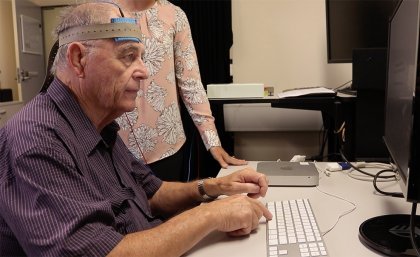 Man sitting at computer with electrical stimulation machine on head with researcher standing over his shoulder.