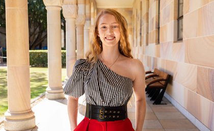 A young woman with long blonde hair standing under a sandstone walkway surrounded by columns