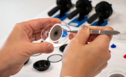 a closeup image of hands holding tweezers and a clear dish containing a paper-like disk with a black mark in its centre 