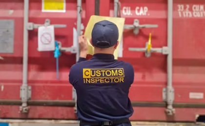a man in a blue shirt with Customs Inspector on it looks at a red shipping container