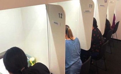 Women sitting behind desks in individual booths with their backs to the camera tasting food