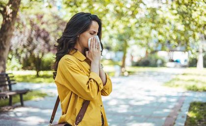 Woman in yellow jacket blowing her nose in a tissue standing in a city park with trees in the background 