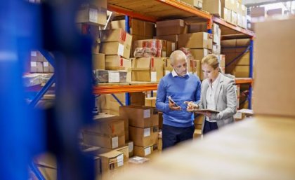 Image: Adobe. A man and a woman look at a clipboard in a factory while surrounded by boxes