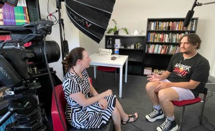 UQ Bachelor of Arts Alum Jamaine Wilesmith was interviewed by Associate Professor Katelyn Barney for the project. Two people sitting in a room facing eachother, surrounded by filming equipment including a camera and light diffuser