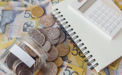 Image: Adobe. A notebook and calculator sit next to bank notes and a jar of coins