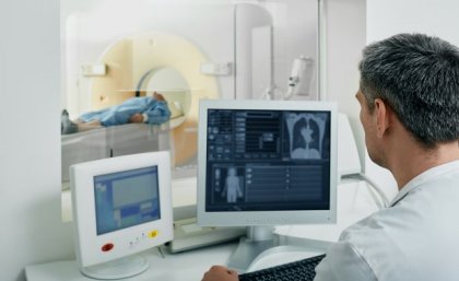 Image, Adobe. Man in white coat looking at radiology images with patient in CT scanner in background. Adobe.