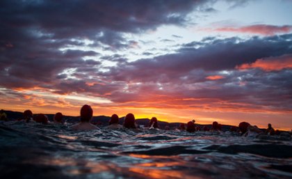 Image: Courtesy Dark Mofo. A sunrise photo taken from the water with swimmers in silhouette against a cloudy orange sky.