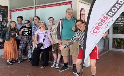 Crossroad Arts is an inclusive arts organisation for people with a disability or who are deaf. Image: Crossroad Arts. A group of people smiling at the camera holding a flag that says "Cross Roads".