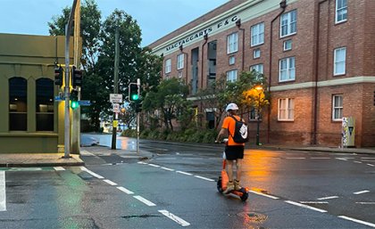 Image: The University of Queensland. A man in high-vis and wearing a helmet and backpack rides an e-scooter across a Brisbane road.