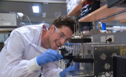 Dr Aiden Beauglehole working with the UQ bioreactors. a man in a white lab coat and glasses adjusts a glass cylinder surrounded by stainless steel equipment