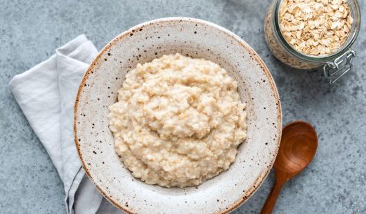 a bowl of porridge sits next to a wooden spoon and a jar of uncooked oats