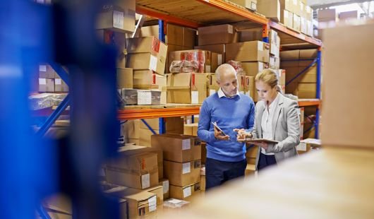 A man and a woman look at a clipboard in a factory while surrounded by boxes