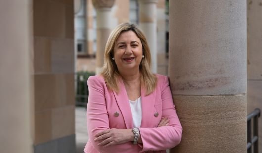 A woman in a pink blazer leans against a sandstone pillar, smiling at the camera. 