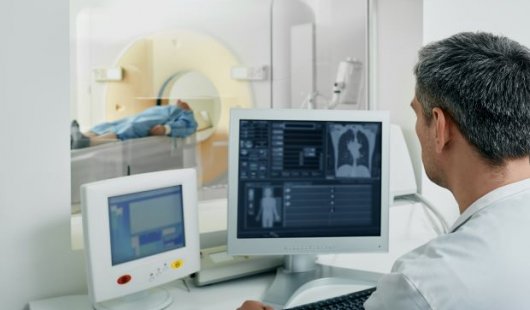 Man in white coat looking at radiology images with patient in CT scanner in background. Adobe.