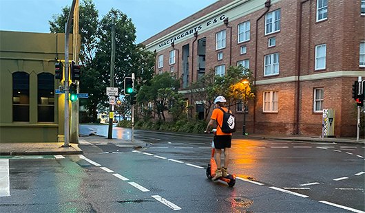 A man in high-vis and wearing a helmet and backpack rides an e-scooter across a Brisbane road.