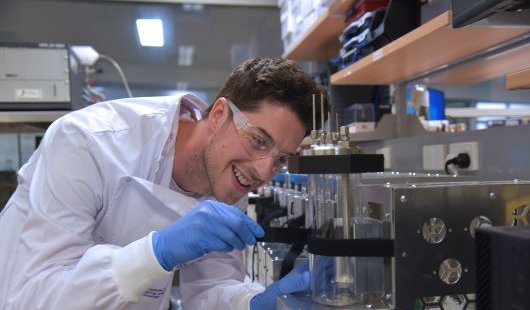 a man in a white lab coat and glasses adjusts a glass cylinder surrounded by stainless steel equipment