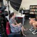 UQ Bachelor of Arts alum Jamaine Wilesmith interviewed by Associate Professor Katelyn Barney for the project. Two people sitting in a room facing eachother, surrounded by filming equipment including a camera and light diffuser