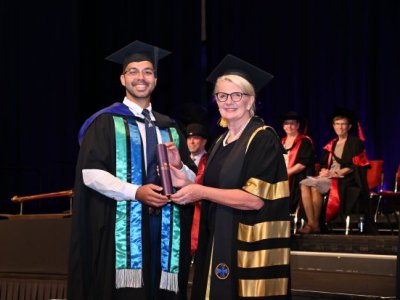 A man and a woman in graduation attire posing on stage - the man holding a cylinder 