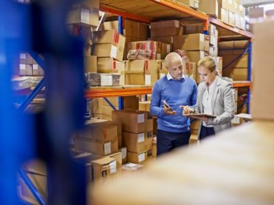 A man and a woman look at a clipboard in a factory while surrounded by boxes