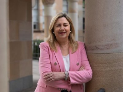 A woman in a pink blazer leans against a sandstone pillar, smiling at the camera. 