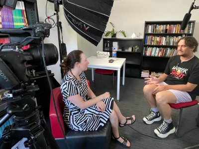 Two people sitting in a room facing eachother, surrounded by filming equipment including a camera and light diffuser 