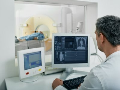 Man in white coat looking at radiology images with patient in CT scanner in background. Adobe.