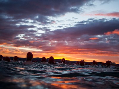 A sunrise photo taken from the water with swimmers in silhouette against a cloudy orange sky.