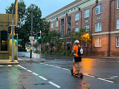 A man in high-vis and wearing a helmet and backpack rides an e-scooter across a Brisbane road.