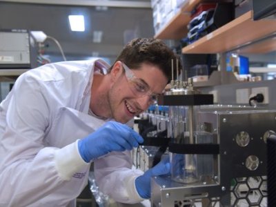 a man in a white lab coat and glasses adjusts a glass cylinder surrounded by stainless steel equipment