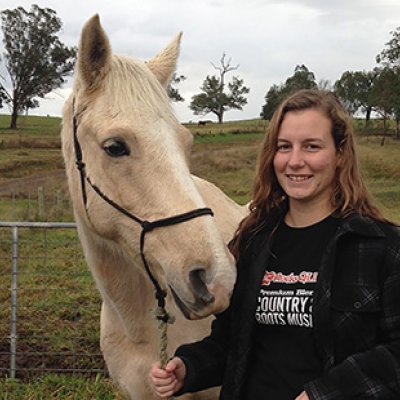 Carissa Horsey, a young woman, standing and smiling next to her light-coloured horse in a field