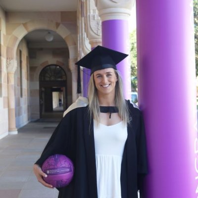 A young woman in a graduation cap and gown holds a netball and leans against a column in the St Lucia Great Court