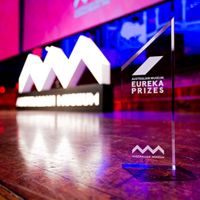 A glass trophy sitting on a wooden floor. Red and purple lights illuminate the room. There is a white sign reading Australian Museum in the background.