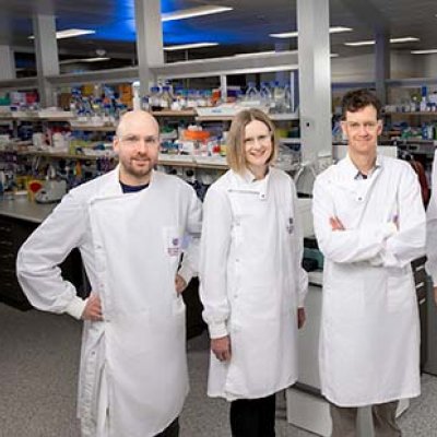 Five scientists wearing UQ-branded lab coats in a laboratory. They are lined up in a row and smiling.