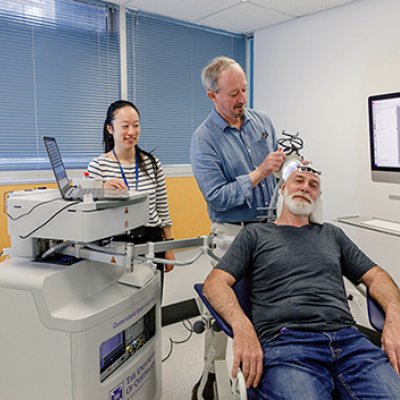 A man sitting on a chair receiving ultrasound treatment on his head in a lab. Two ultrasound technicians are behind him.