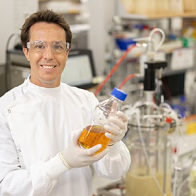 Associate Professor Tim Mercer wearing a lab coat and safety goggles holding a bottle containing orange liquid