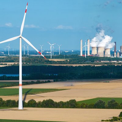 A wind turbine in a dry field with a power plant emitting steam in the background