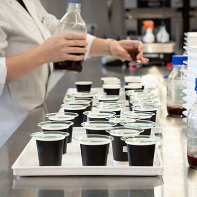 Small coffee cups on a bench in a lab. A person wearing a lab coat is holding a bottle of coffee.