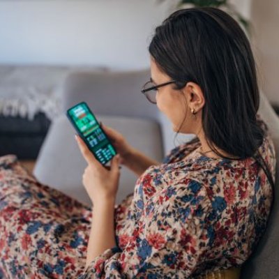 A woman sits on a couch looking at a phone showing a sports betting app