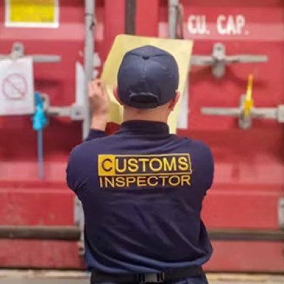 a man in a blue shirt with Customs Inspector on it looks at a red shipping container