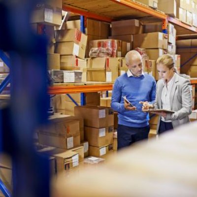 A man and a woman look at a clipboard in a factory while surrounded by boxes